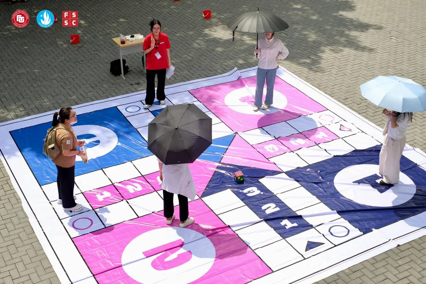 A giant board game laid out on the forecourt, players under umbrellas